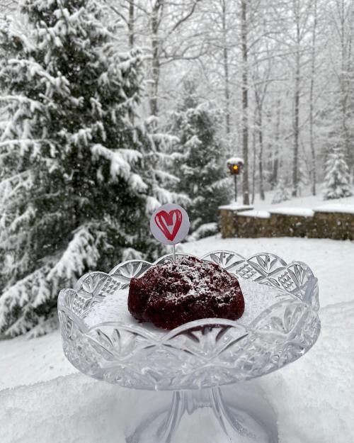 A red velvet dessert on a crystal plate, with a heart-shaped decoration, set against a snowy forest backdrop.
