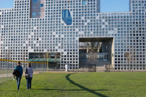 Two people walk towards a modern building with a unique facade of square windows, set against a grassy area.