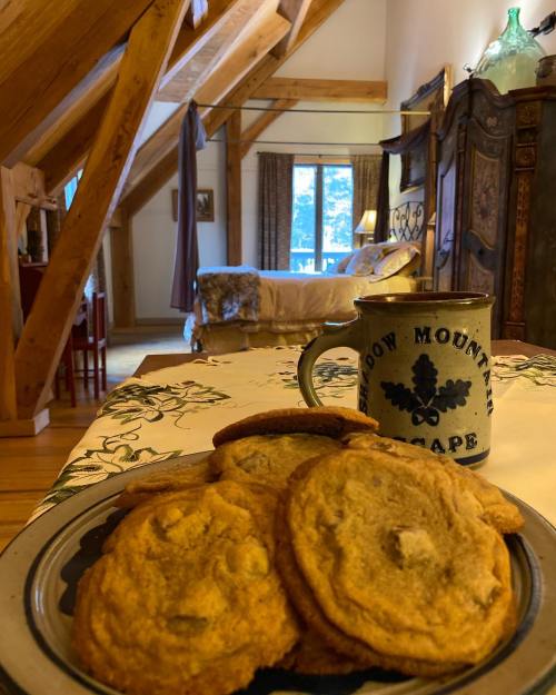 A cozy cabin interior with a plate of cookies and a mug on a table, featuring wooden beams and a view of a bedroom.