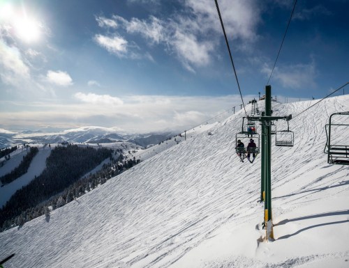 Two skiers on a chairlift ascend a snowy mountain under a bright blue sky with scattered clouds.