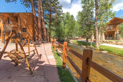 A peaceful outdoor scene with a wooden swing, a stream, and trees beside rustic buildings under a blue sky.
