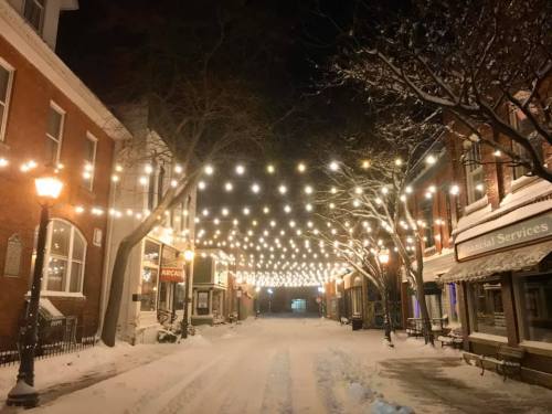 A snowy street at night, lined with buildings and illuminated by string lights overhead.