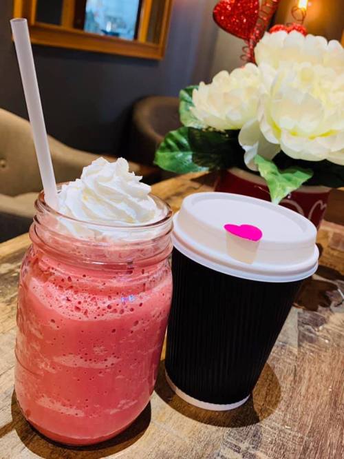 A pink milkshake topped with whipped cream next to a black coffee cup with a heart-shaped lid, on a wooden table.