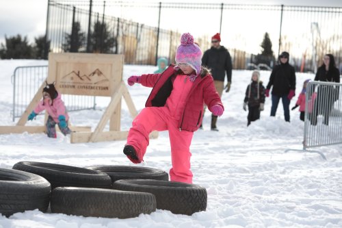 A child in a pink snowsuit steps on tires in a snowy outdoor setting, with other children and adults in the background.
