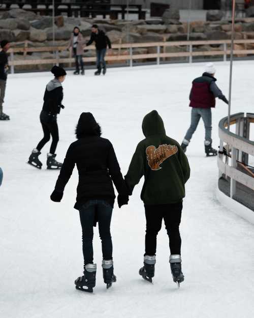 Two people skate hand in hand on an ice rink, surrounded by others enjoying the activity.