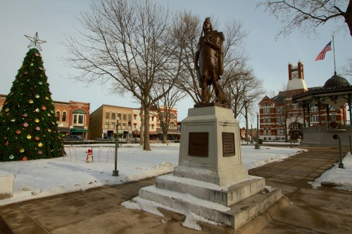 A winter scene featuring a statue, a decorated Christmas tree, and a historic building in a snowy town square.