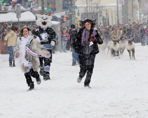 Two women run through a snowy street, one in a fur coat, alongside a mascot and reindeer in a festive winter event.
