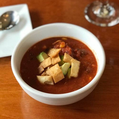 A bowl of soup topped with tortilla strips and avocado, placed on a wooden table. A spoon and glass are in the background.
