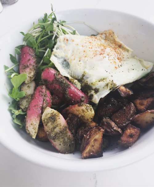 A bowl with roasted potatoes, sautéed radishes, microgreens, and a fried egg, garnished with herbs and spices.