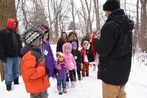 A group of children in winter clothing listens to an adult in the snow, surrounded by trees. Snow is falling gently.