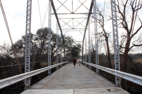 A person walks on a metal bridge surrounded by trees and a rural landscape under a cloudy sky.