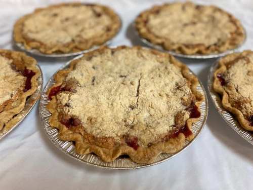 Five freshly baked fruit pies with crumbly tops, arranged on a white tablecloth.