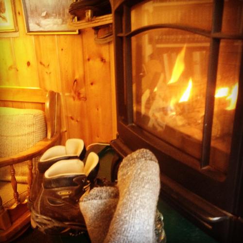 Cozy scene with feet in socks resting near a warm fireplace, with a pair of boots nearby in a rustic cabin setting.