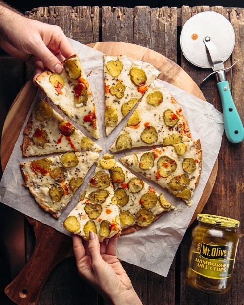 A pizza topped with pickles and peppers, sliced into pieces, with hands reaching for a slice and a jar of dill chips nearby.