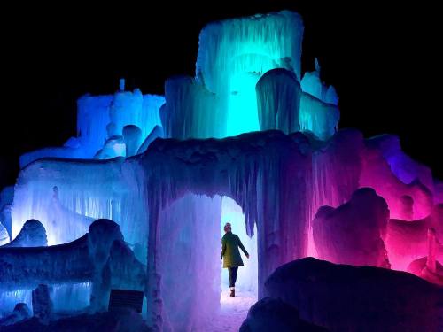 A person walks through a colorful ice structure illuminated in blue, green, and pink lights at night.