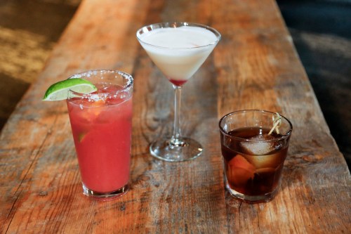 Three cocktails on a wooden table: a pink drink with lime, a martini, and a dark drink with ice and a garnish.