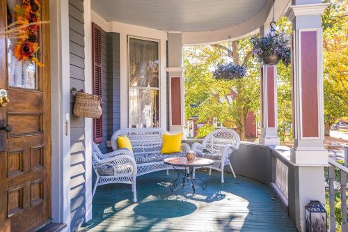 A cozy porch with a wicker sofa, yellow pillow, and a small table, surrounded by autumn foliage and hanging plants.