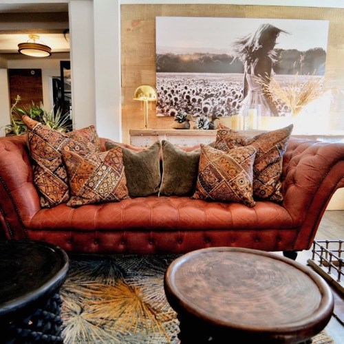 A cozy living room featuring a brown tufted sofa with patterned pillows, wooden tables, and a large wall art piece.
