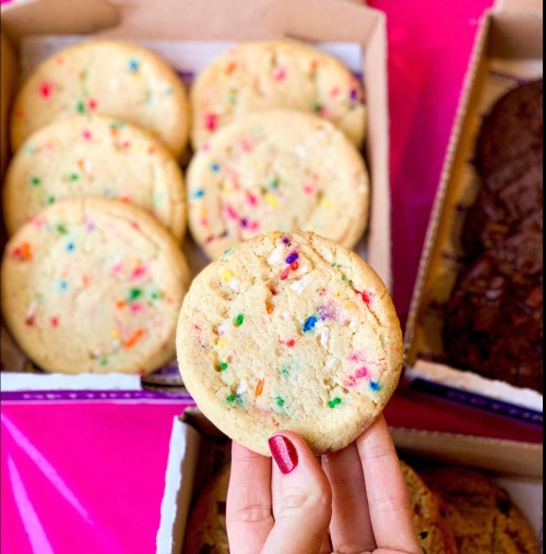 A hand holds a colorful cookie with sprinkles, surrounded by more cookies in boxes on a bright pink surface.
