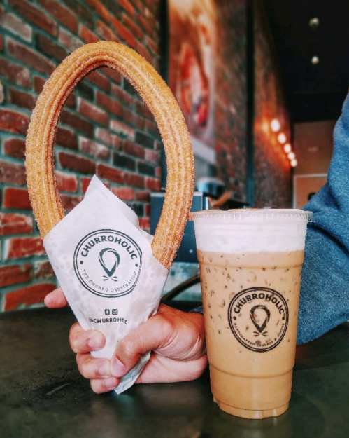 A hand holds a large churro in a paper wrapper next to a cup of iced coffee on a table with a brick wall background.