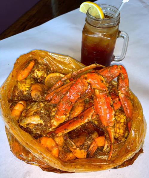 A seafood boil with crabs, shrimp, and corn, served in a bag alongside a glass of iced tea with a lemon slice.