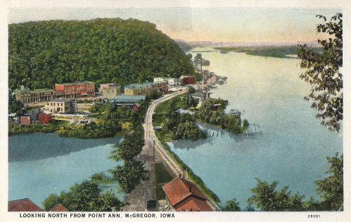 Aerial view of McGregor, Iowa, showcasing the river, buildings, and lush hills in the background during sunset.