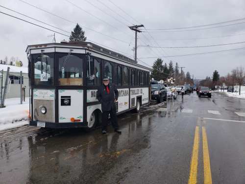A man in a black coat stands next to a vintage trolley bus on a snowy street, with vehicles and trees in the background.