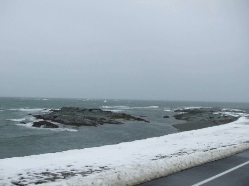 A gray, overcast day at the coast, with rocky shores and patches of snow along the water's edge.