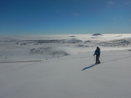 A person stands on a snowy landscape, overlooking a misty valley and distant hills under a clear blue sky.