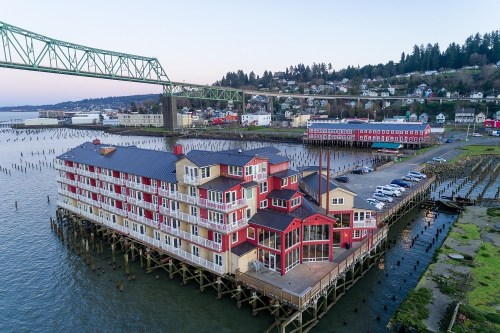 Aerial view of a red and white building on stilts by the water, with a bridge and town in the background.