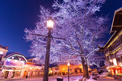 A snowy street scene at night, featuring a tree adorned with lights and festive storefronts in a charming village.