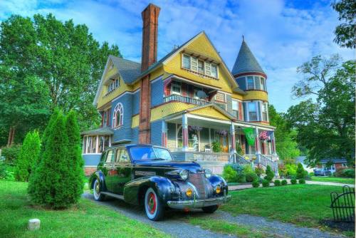 A vintage black car parked in front of a colorful Victorian-style mansion surrounded by lush greenery.
