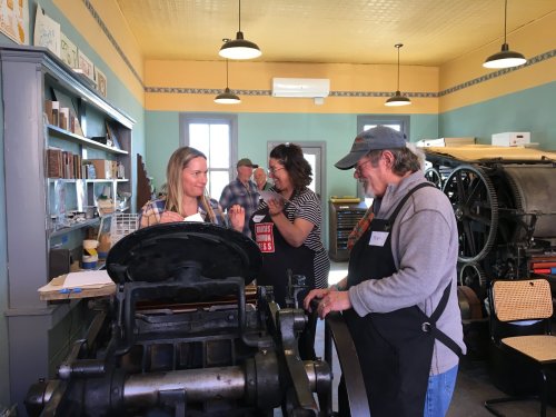 Two women and a man engage in conversation near a vintage printing press in a colorful workshop setting.