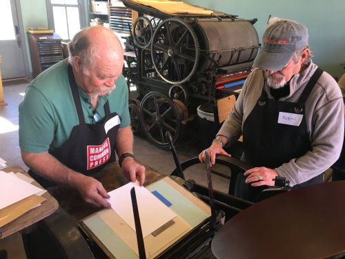 Two people work together at a printing press, preparing paper for printing in a workshop setting.