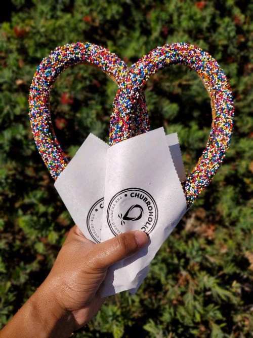 A hand holds two large, colorful, sprinkle-covered churros in paper wrappers against a green background.