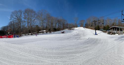 A snowy ski slope with tracks, trees in the background, and a ski lift on a clear blue sky day.