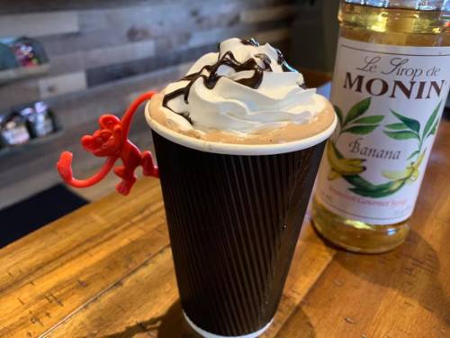 A cup of chocolate drink topped with whipped cream and chocolate drizzle, featuring a red toy figure and a bottle of banana syrup.