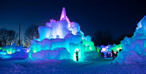 A colorful ice castle illuminated with blue, green, and purple lights against a dark evening sky, with people nearby.