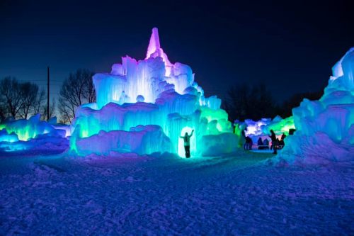 A colorful ice castle illuminated with blue, green, and purple lights against a dark evening sky, with people nearby.
