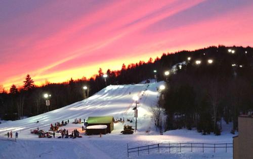 A snowy hill at sunset, with people sledding and a lit-up lodge in the foreground against a vibrant sky.