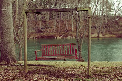 A red wooden swing hangs from a rustic frame by a calm river, surrounded by bare trees and fallen leaves.