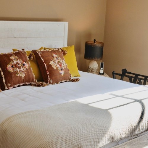 Cozy bedroom with a white bed, decorative pillows, a lamp, and warm-toned walls. Sunlight casts shadows on the bed.