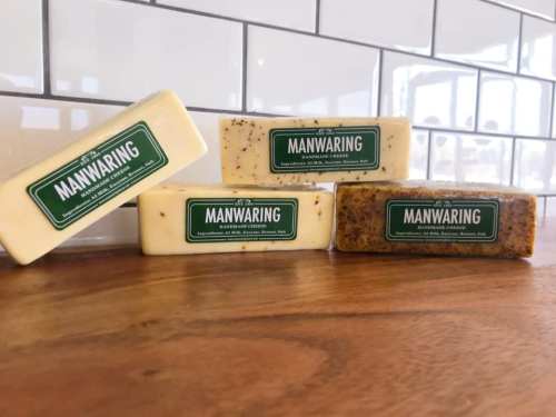 Four bars of artisanal soap with green labels reading "MANWARING," displayed on a wooden surface against a tiled background.
