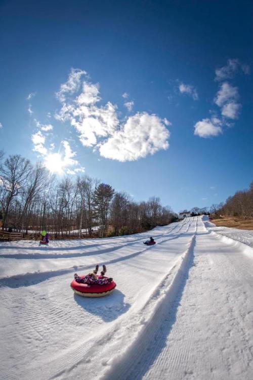 People tubing down a snowy slope under a bright blue sky with fluffy clouds. Trees line the background.