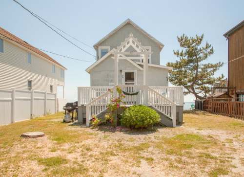 A charming beach house with a front porch, surrounded by greenery and a sandy yard, near the ocean.