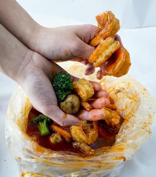 Hands holding shrimp, broccoli, and other seafood in a spicy sauce, with a messy, colorful background.