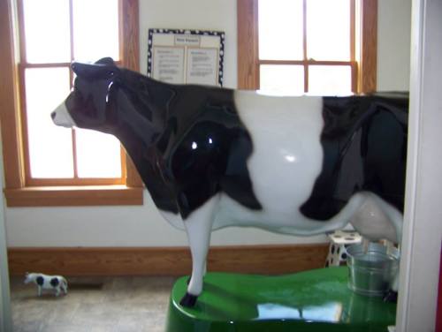 A life-sized black and white cow statue stands in a room with large windows and a small toy cow nearby.