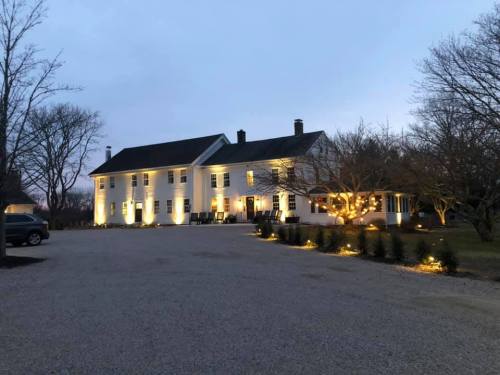 A well-lit white house at dusk, surrounded by trees and a gravel driveway, creating a warm and inviting atmosphere.