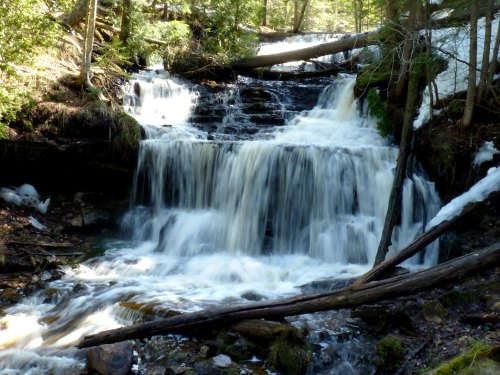 A serene waterfall cascades over rocks, surrounded by lush greenery and remnants of snow in a tranquil forest setting.