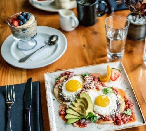 A plate of eggs with avocado and salsa, alongside a glass of water and a dessert with fruit and oatmeal.